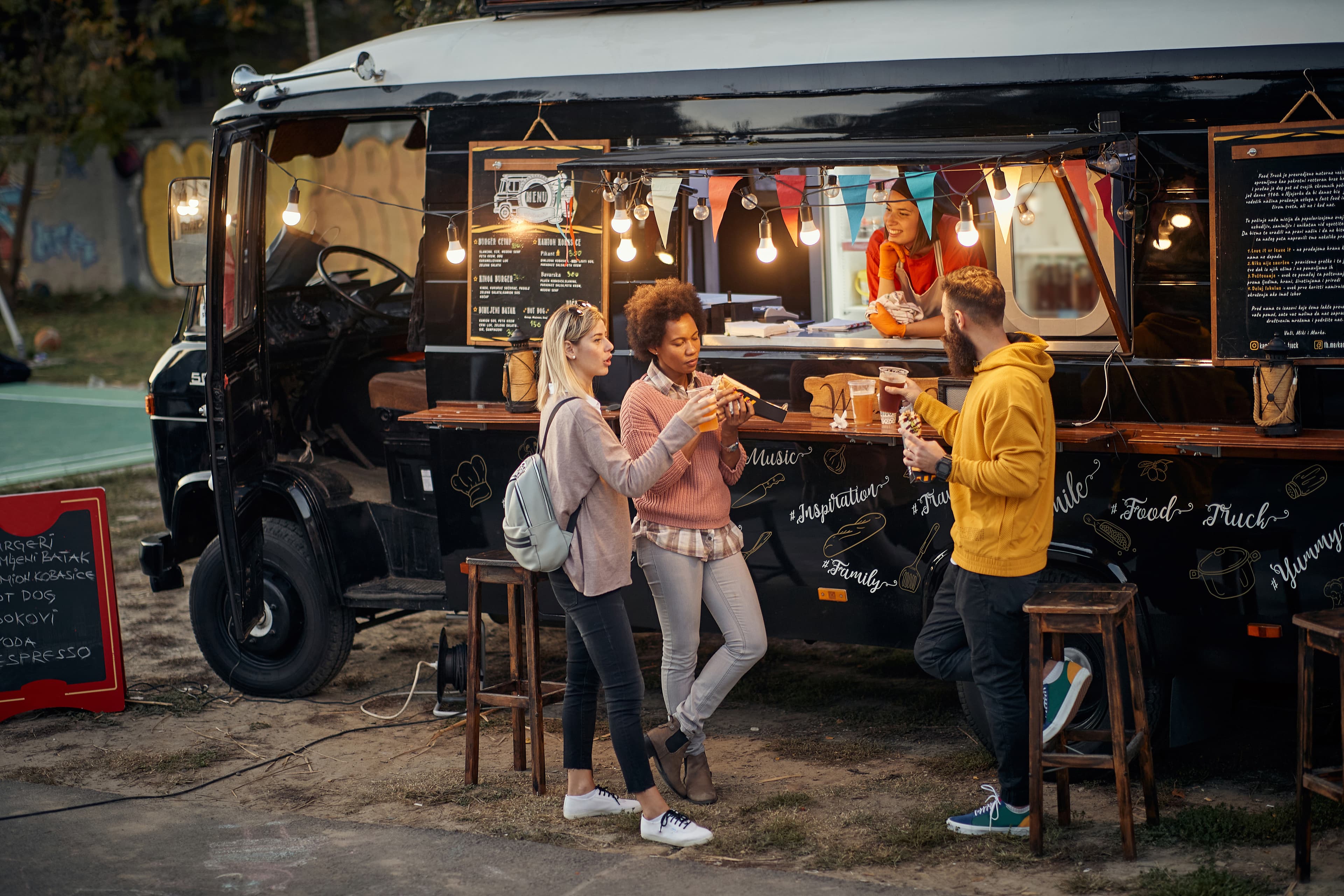 two women and a man stand in front of a black food truck enjoying food and drink served by a woman in a red shirt. the food truck has a menu on the side 